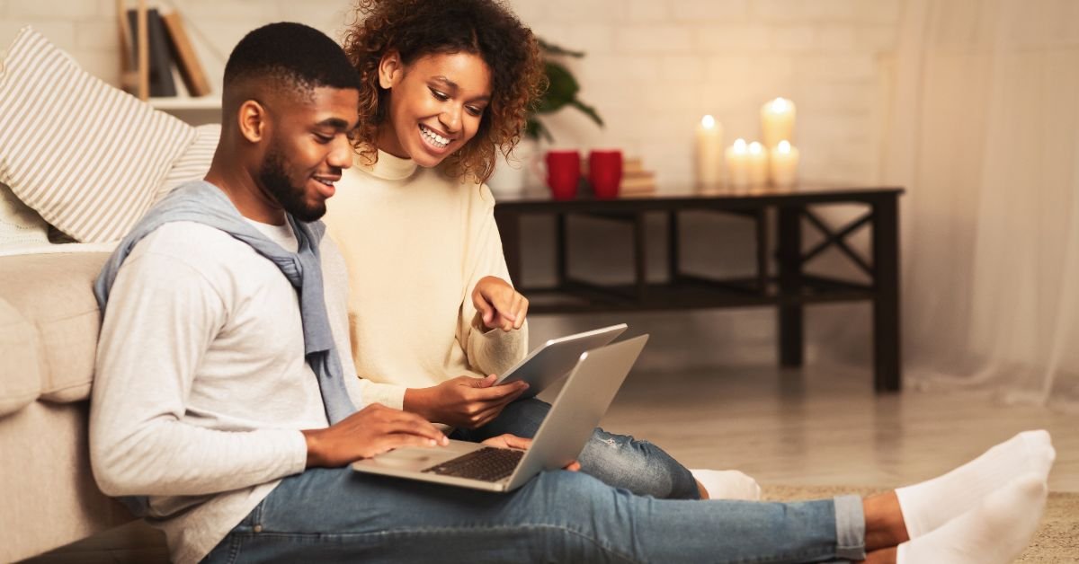 A young couple sits on the floor, focused on a laptop, sharing ideas and enjoying each other's company.