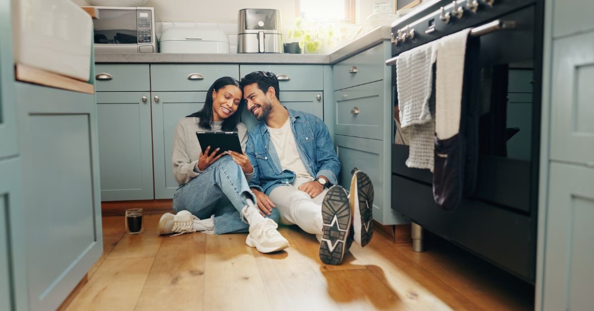 A man and woman are seated on the kitchen floor, enjoying each other's company amidst a cozy kitchen setting.
