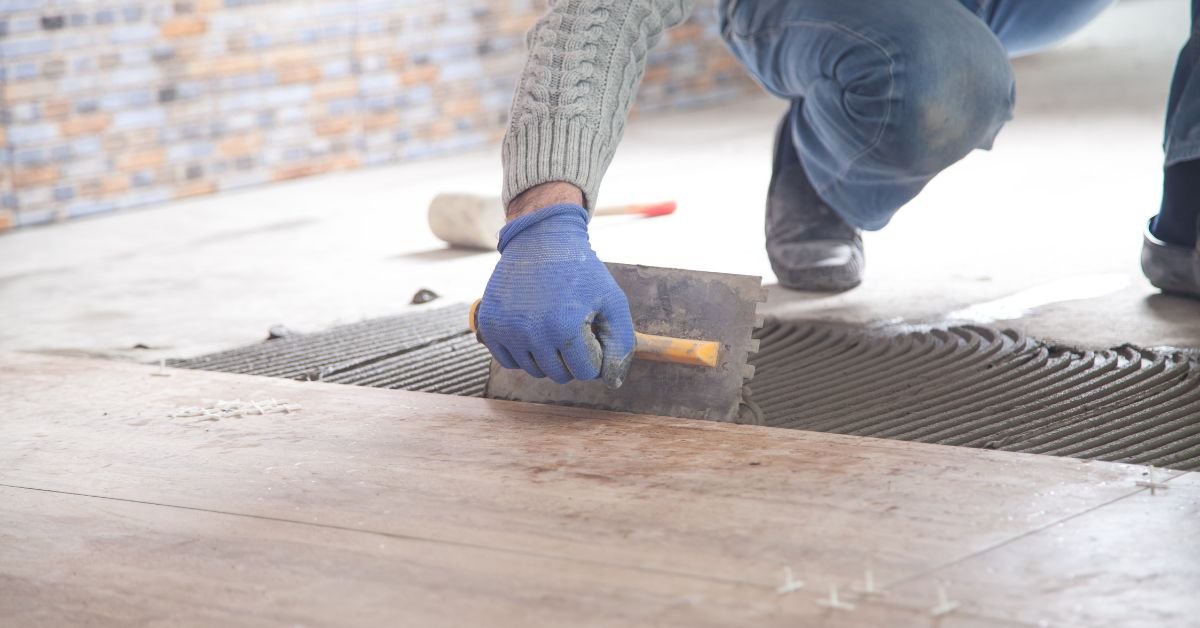 A man is positioned on the floor, applying adhesive and placing tiles in a precise arrangement.