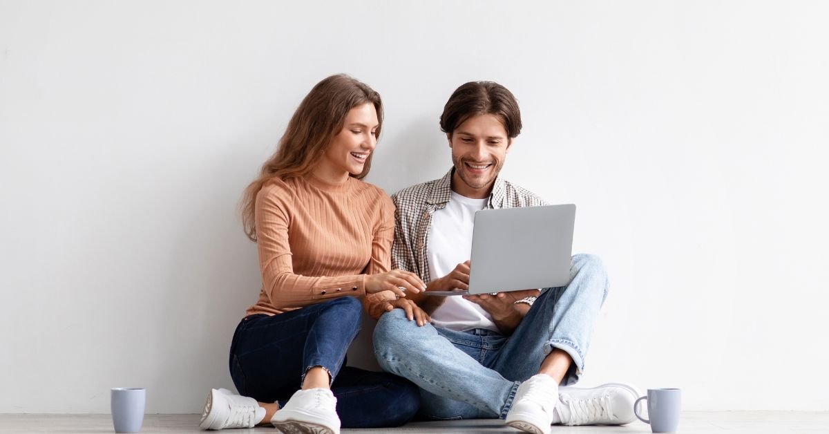A man and woman are seated on the floor, using a laptop together, appearing to share ideas or work on a project.
