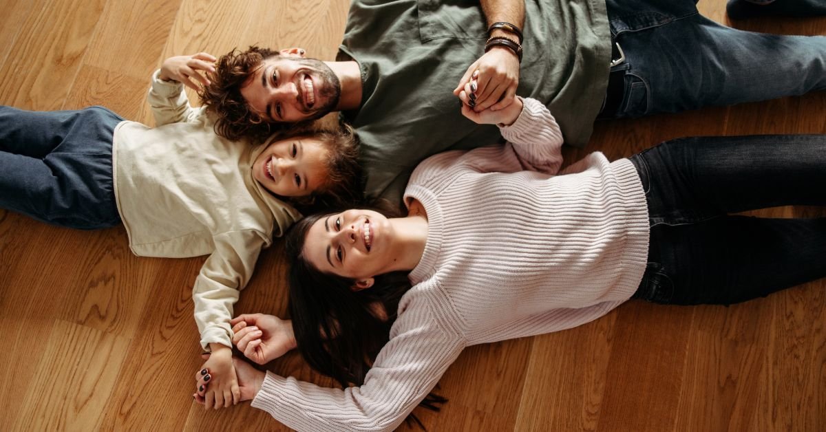 A family of four, including two adults and two children, lying together on the floor in a cozy living room setting.