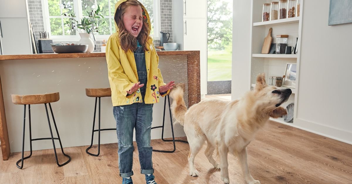 In a kitchen, a girl wearing a yellow raincoat stands next to a dog, both appearing cheerful and engaged.