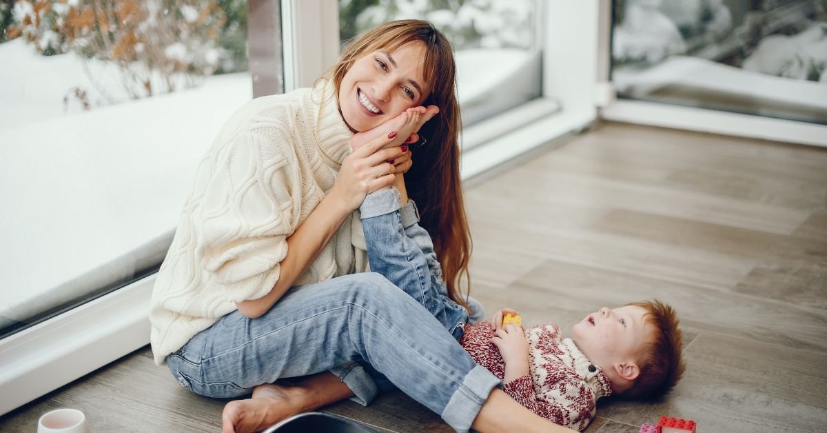 A woman sits on the floor with a baby in her lap, both engaged in a playful moment together.