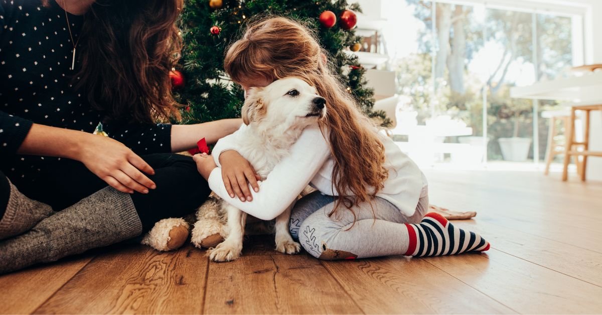 A woman and child sit on the floor, playfully interacting with a dog beside them.